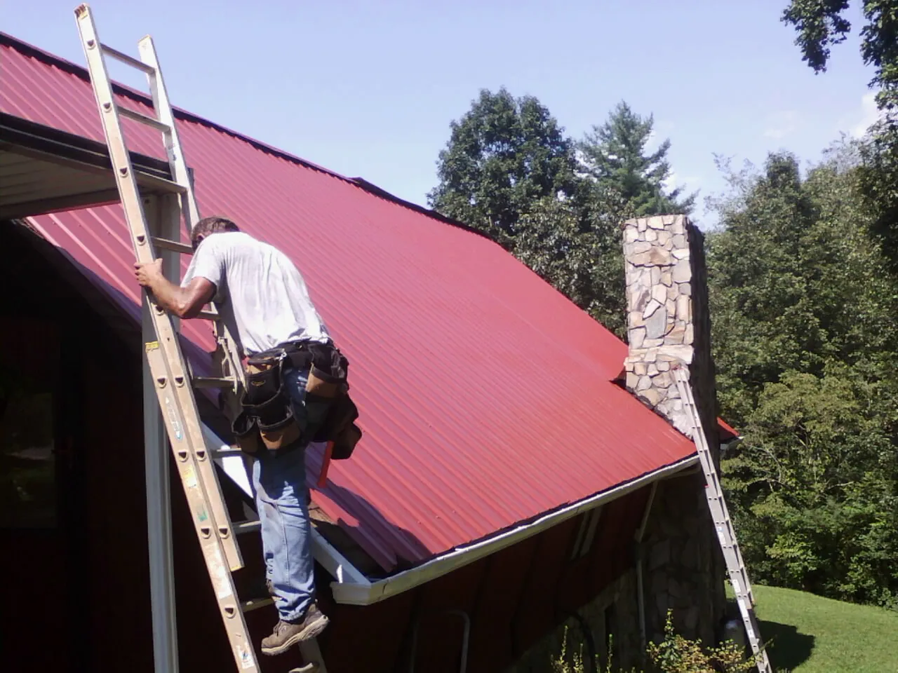 Roofer on a ladder beside a red metal roof and stone chimney — Soffit & Fascia Repair by Roofing Brothers in Hood